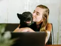 Mujer besa a su perro velcro, un labrador negro, mientras trabaja en su portátil.